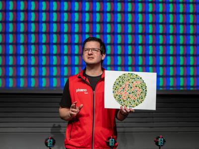 Mann hält ein Bild in der Hand, darauf ist zwischen grünen Punkten eine Zahl aus roten Punkt abgebildet.