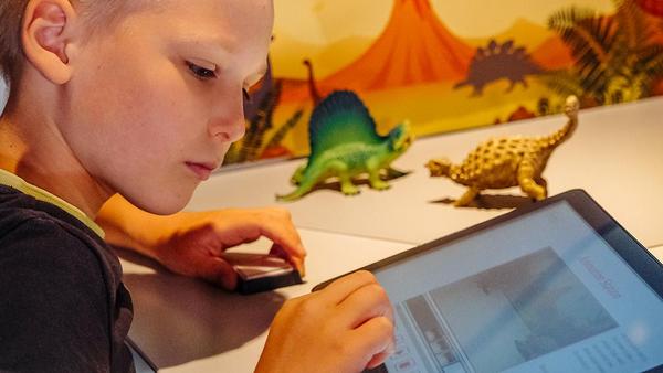 Junge arbeitet an einem Tablet. Im Hintergrund sieht man Dinosaurierfiguren